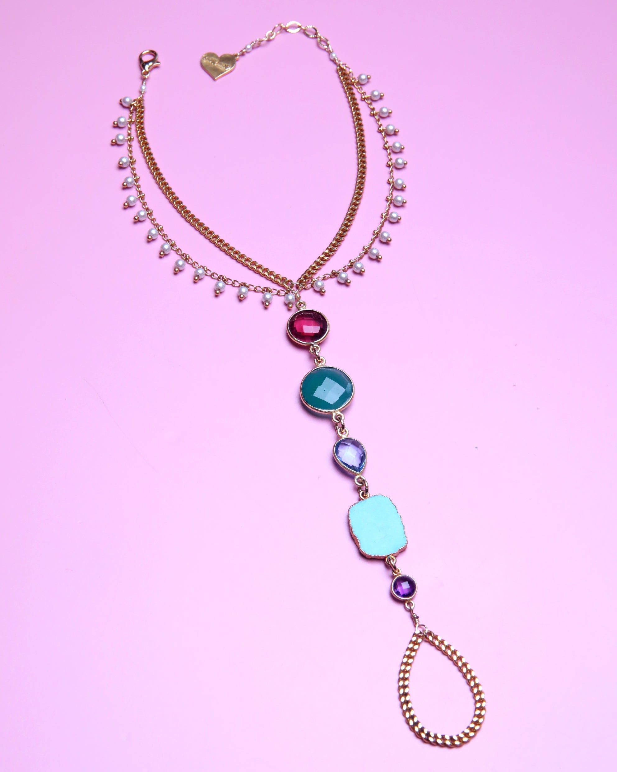 A gold-toned double-strand necklace with a long drop chain featuring colorful faux gemstones in pink, teal, light blue, and purple, displayed on a solid pink background.