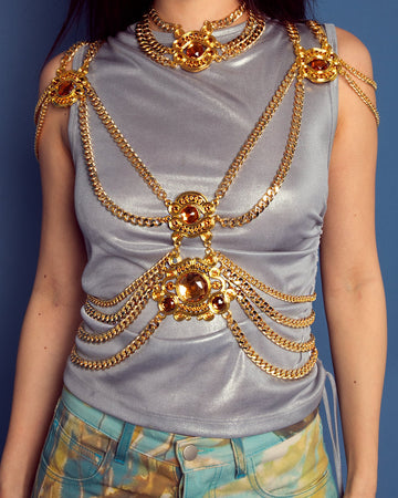 Woman wearing a silver top with gold chains and patterned pants against a blue background